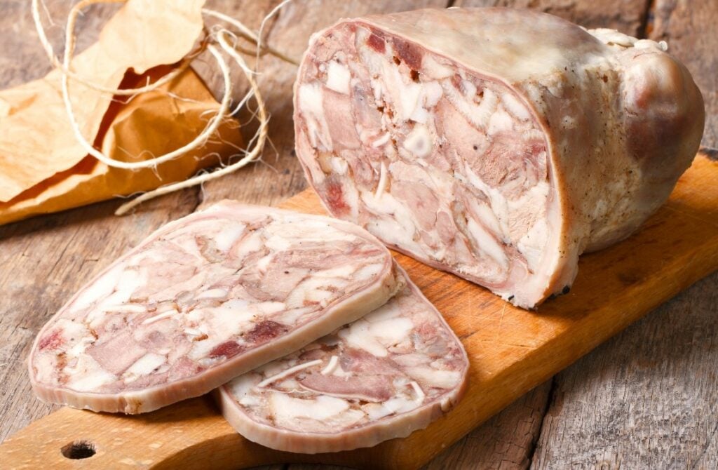 A loaf of head cheese with two slices cut and placed on a wooden cutting board; twine and parchment are in the background on a rustic wooden surface.