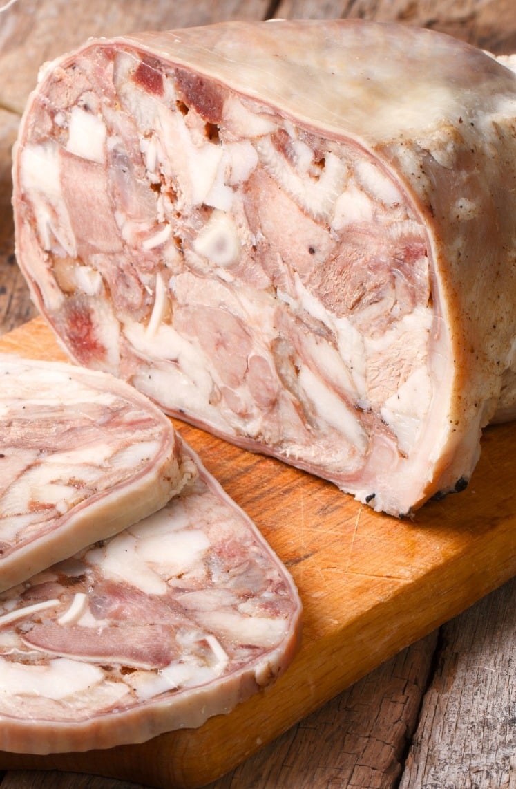 A loaf of head cheese with two slices cut, showing a mosaic of meat and fat, sits on a wooden cutting board atop a rustic wooden table.