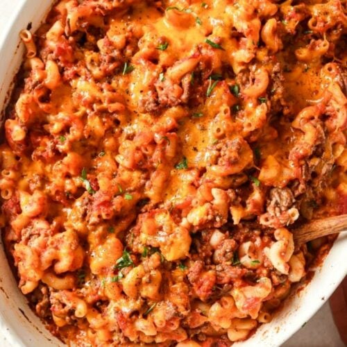 A close-up of a baked casserole dish filled with cheesy, saucy ground beef and macaroni, topped with melted cheddar cheese. A wooden spoon is partially visible in the dish.