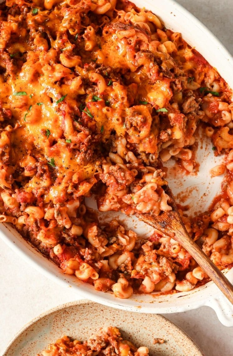 A close-up of baked macaroni with ground beef, tomato sauce, and melted cheese in a white dish, with a wooden spoon serving a portion onto a plate below.
