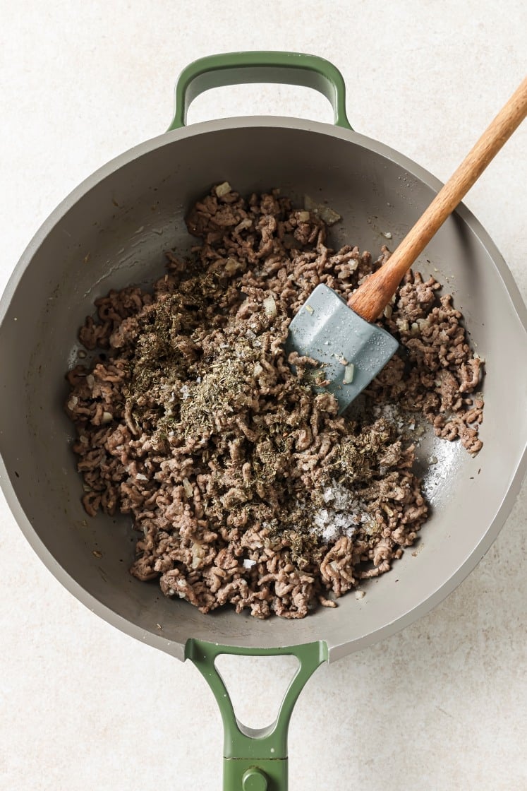 Ground beef is being cooked in a green-handled skillet, seasoned with herbs and spices. A spatula rests in the pan, stirring the mixture as it browns. The background is a light, off-white surface.