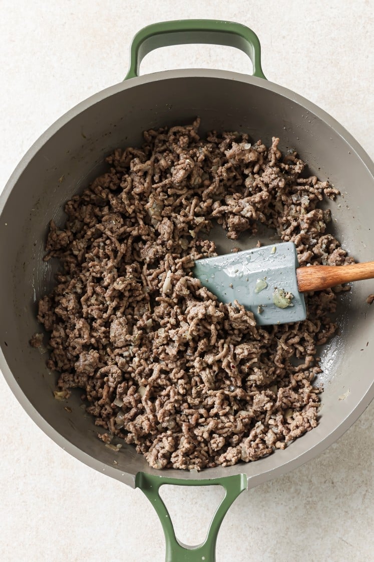 Cooked ground beef in a large gray pan with green handles, being stirred with a gray spatula that has a wooden handle. The beef is browned and crumbled.