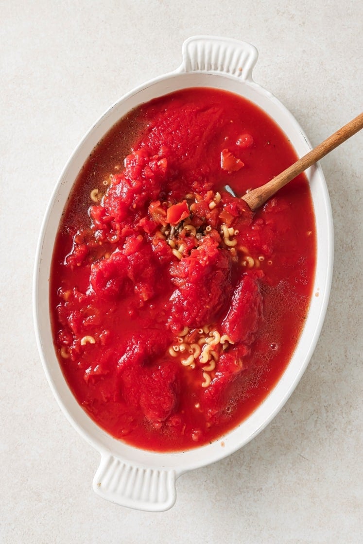 Oval white dish with elbow macaroni, crushed tomatoes, and liquid, being stirred with a wooden spoon, viewed from above on a light surface.