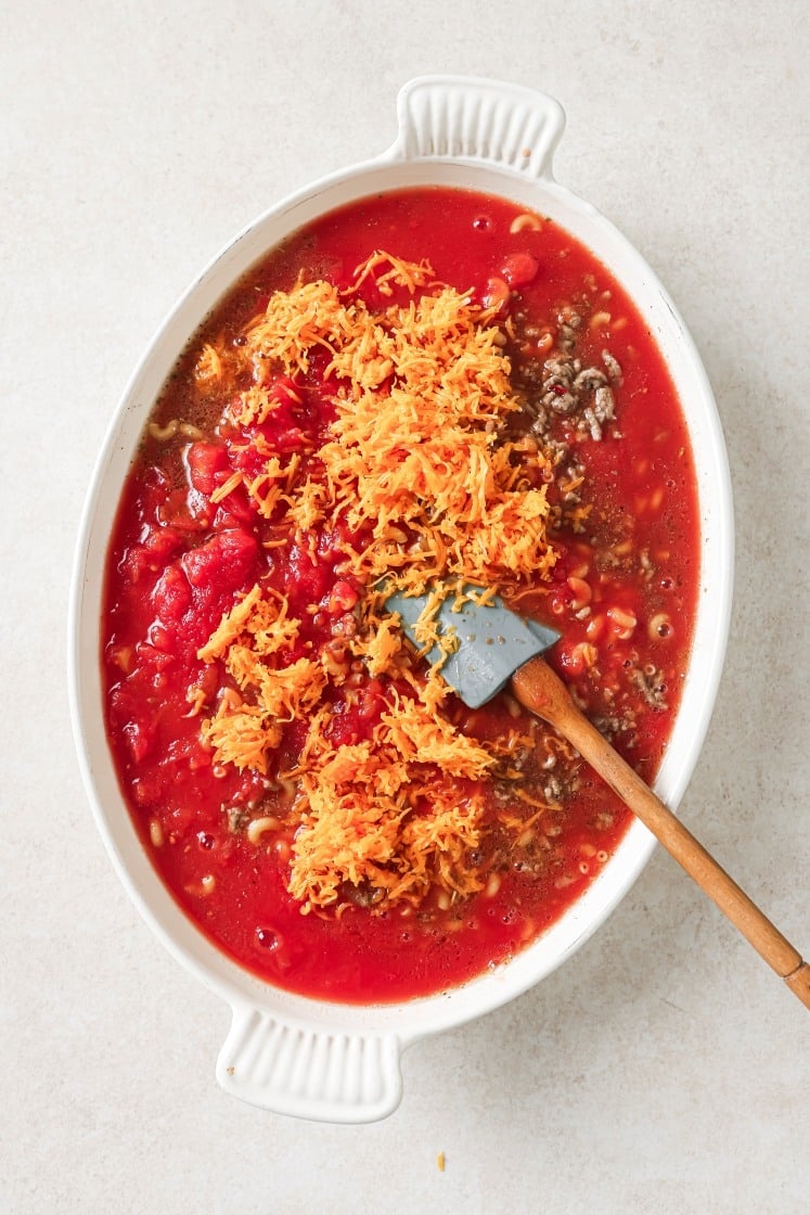 An oval white baking dish filled with ground meat, diced tomatoes, broth, and a mound of shredded orange cheese, being stirred with a spatula on a light countertop.