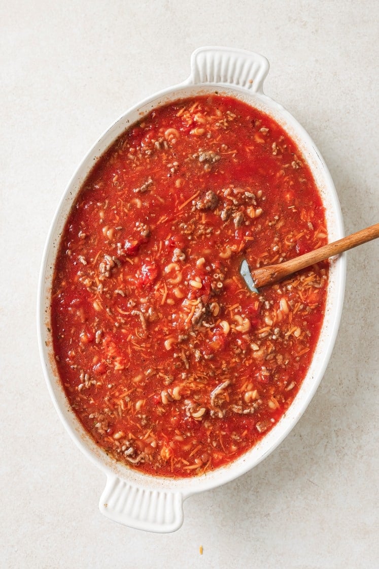 An oval white dish filled with a chunky red soup or stew containing ground beef, tomatoes, macaroni, and possibly shredded cheese, with a wooden spoon resting inside.
