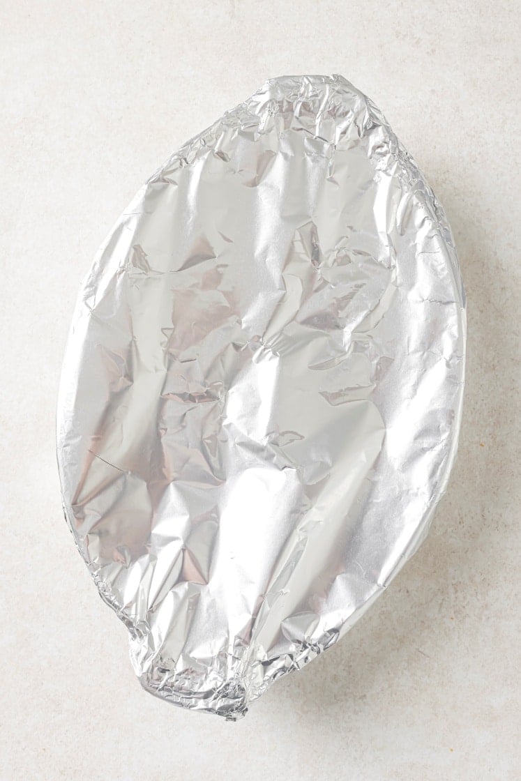 An oval baking dish covered tightly with a sheet of crinkled aluminum foil, placed on a light-colored surface.