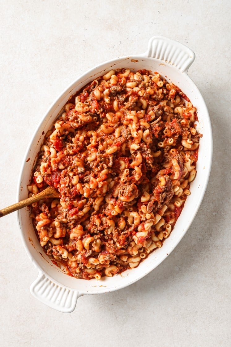 An oval white casserole dish filled with cooked elbow macaroni mixed with ground meat and tomato sauce, with a wooden spoon resting inside.