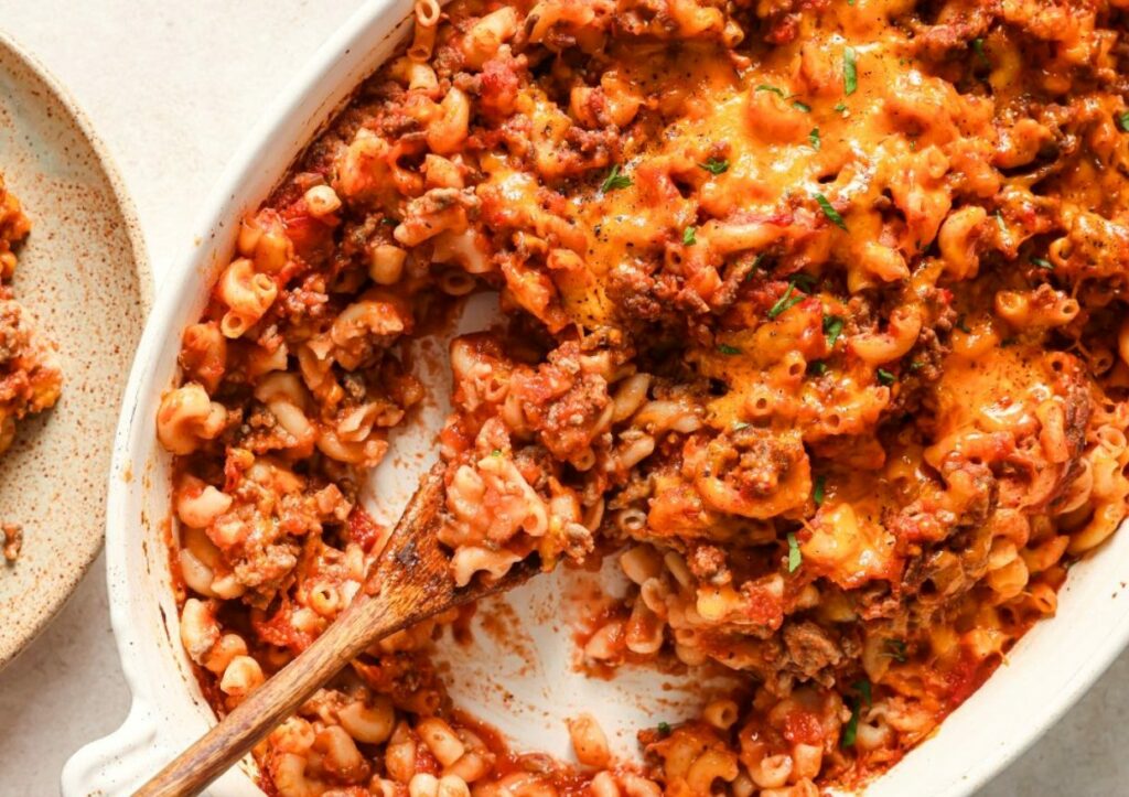 A baked pasta casserole with elbow macaroni, ground meat, tomato sauce, and melted cheese, with a wooden spoon scooping some out. The dish is served in a white baking dish on a light surface.