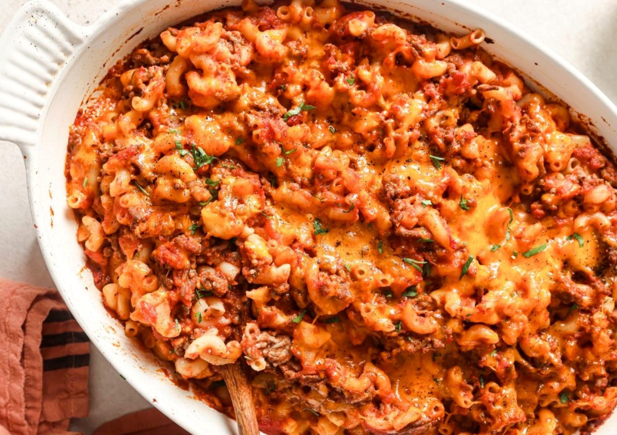 A close-up of a baked casserole dish filled with cheesy, saucy ground beef and macaroni, topped with melted cheddar cheese. A wooden spoon is partially visible in the dish.