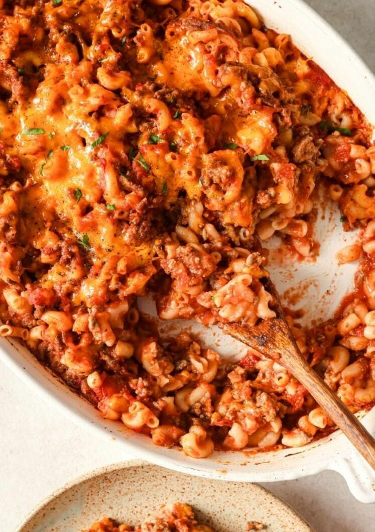 A close-up of a baked macaroni casserole with ground beef, tomato sauce, and melted cheese in a white dish. A wooden spoon is scooping some of the cheesy pasta mixture.
