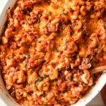 A baked casserole dish filled with cheesy, tomato-based macaroni pasta mixed with ground meat and topped with melted cheese, with a wooden spoon and a folded cloth nearby.