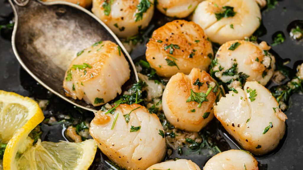 Close-up of golden-brown seared scallops in a skillet with garlic, parsley, and lemon slices, garnished with herbs and glistening with butter.