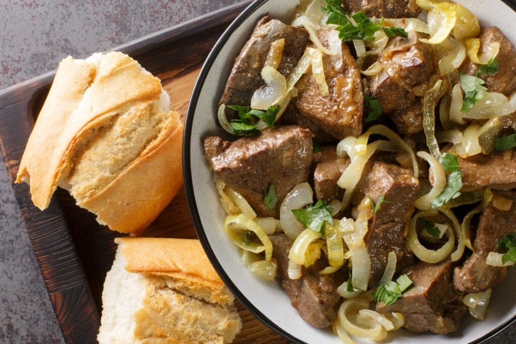 A bowl of cooked liver and onions garnished with parsley, served on a wooden tray next to two pieces of sliced baguette bread.