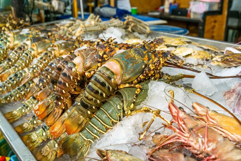 A display of fresh lobsters and large prawns on ice at a seafood market, with more seafood items visible in the background.