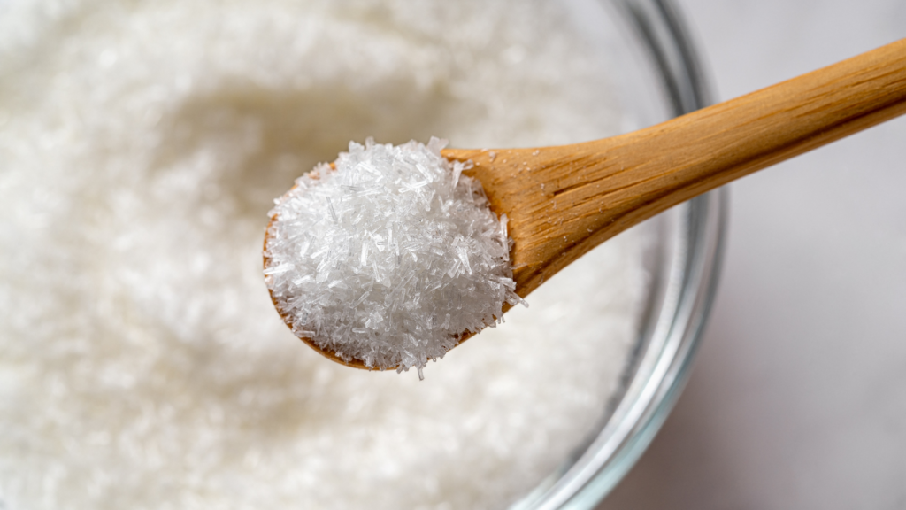 Wooden spoon holding a mound of white, crystalline monosodium glutamate (MSG) granules above a bowl.