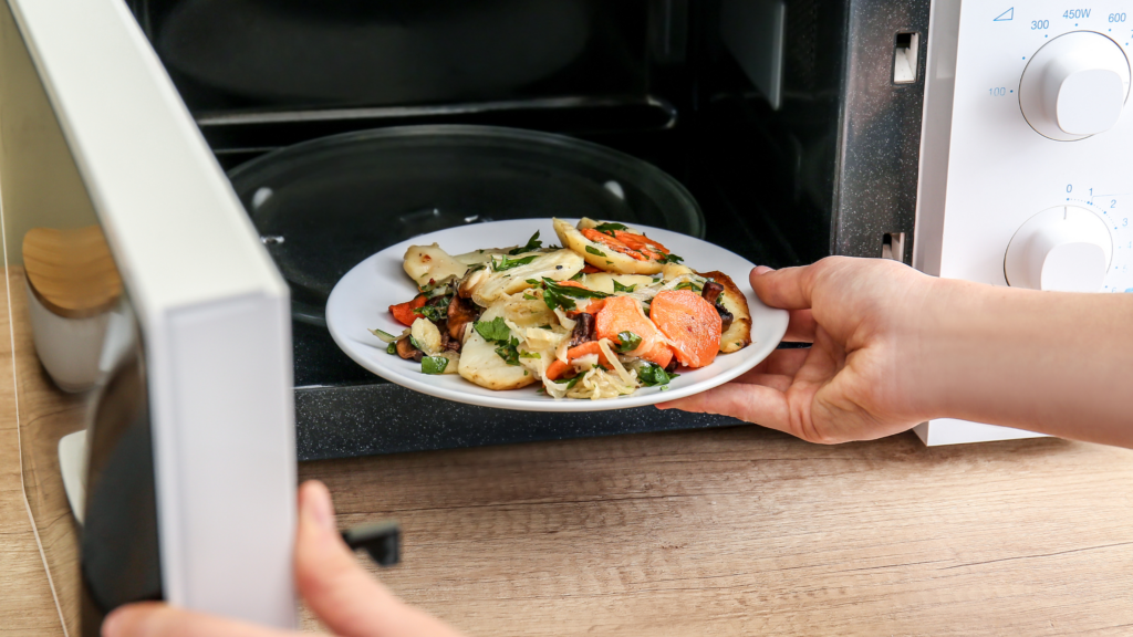 Hands placing a plate of cooked vegetables and potatoes into a microwave with the door open.
