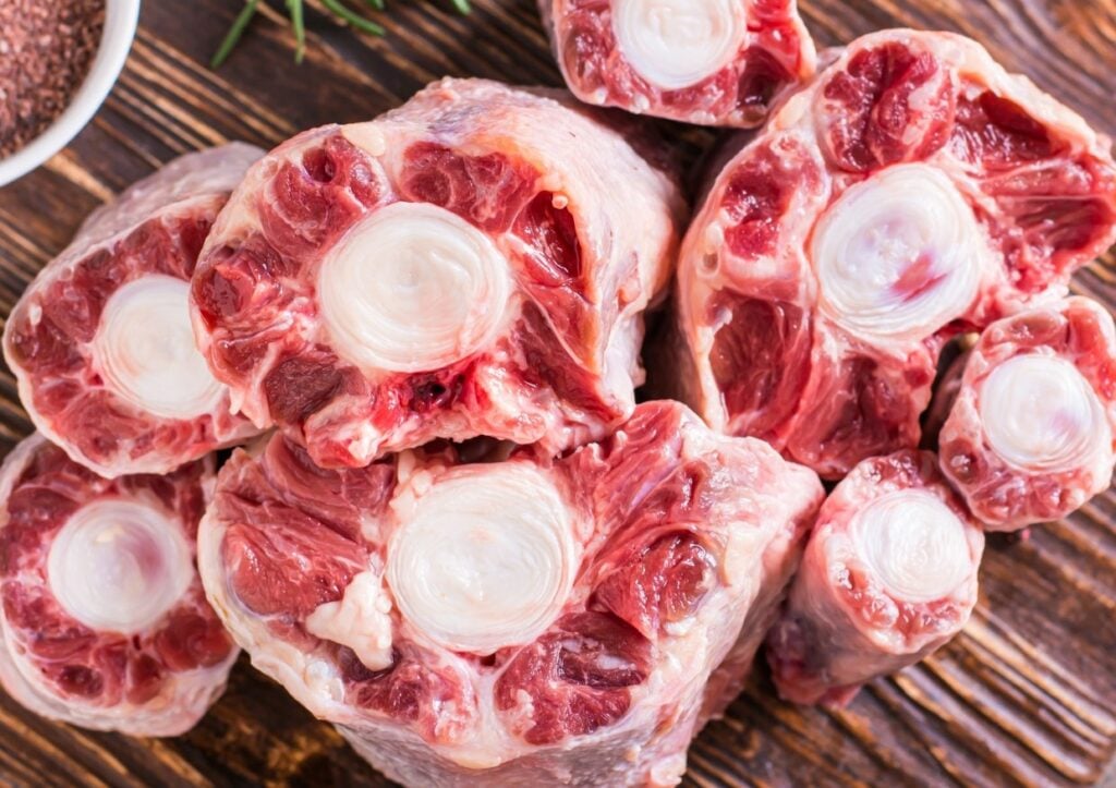 Raw oxtail pieces are arranged on a wooden surface, showing cross-sections of bone surrounded by red meat and white marbling. The texture and details of the meat and bone are clearly visible.