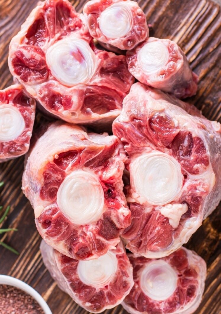 Raw oxtail pieces with visible bone centers and marbled red meat are arranged on a wooden surface. The cuts are circular and show the white bone surrounded by rich, pinkish-red muscle.