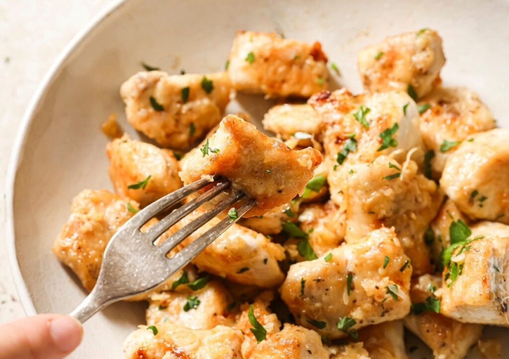 A fork holds a piece of golden-brown, seared chicken garnished with chopped herbs, above a plate filled with similar chicken pieces.