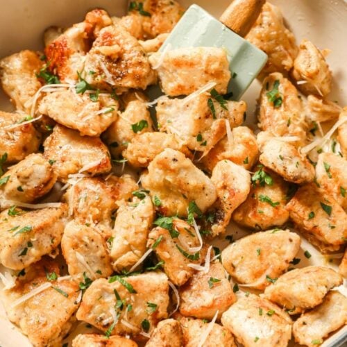 Golden-brown sautéed chicken pieces topped with shredded cheese and chopped herbs in a white skillet, being stirred with a light green spatula.