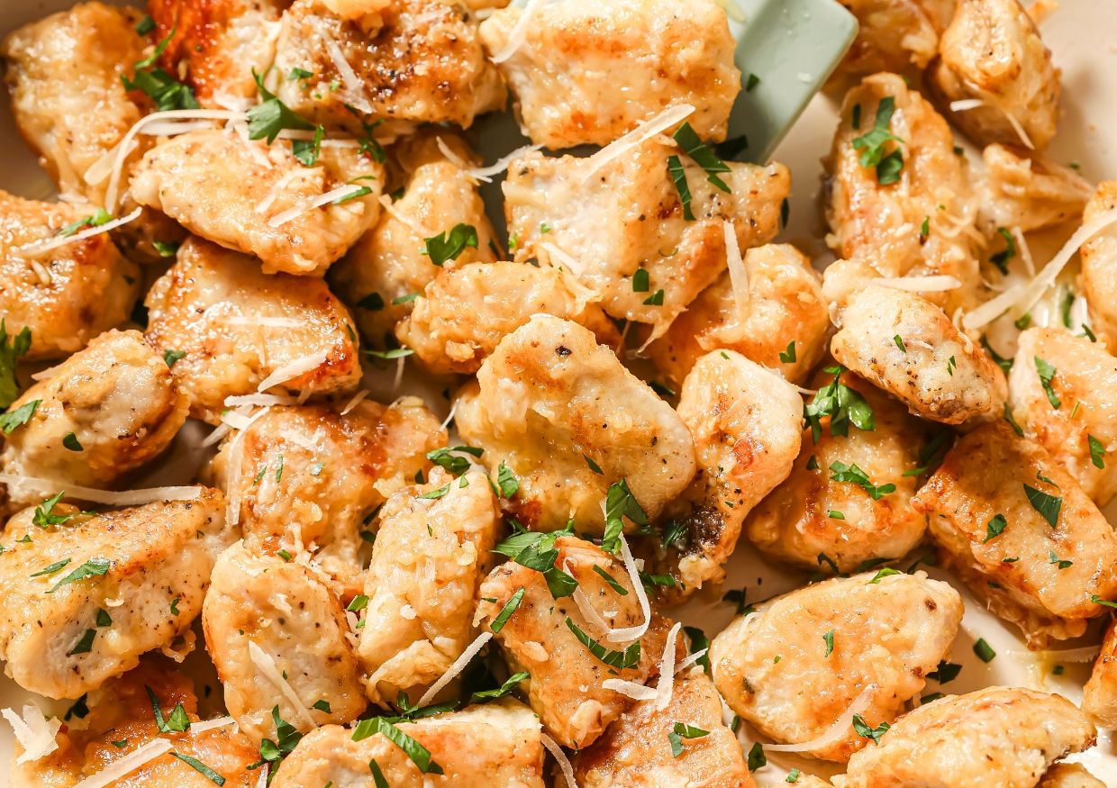 Golden, bite-sized pieces of cooked chicken garnished with chopped parsley and shredded cheese, served on a white plate. The chicken looks crispy and flavorful.
