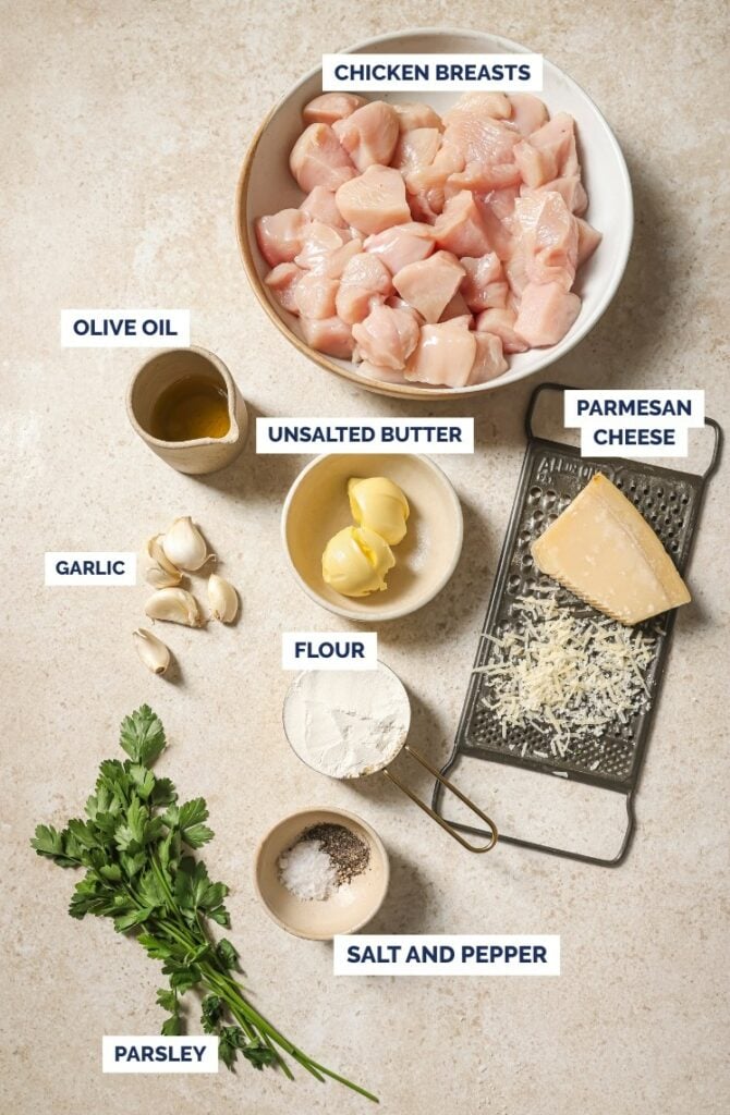 Top-down view of labeled ingredients on a light surface: cubed chicken breasts in a bowl, olive oil, garlic cloves, unsalted butter, flour, grated Parmesan cheese, salt and pepper, and fresh parsley.