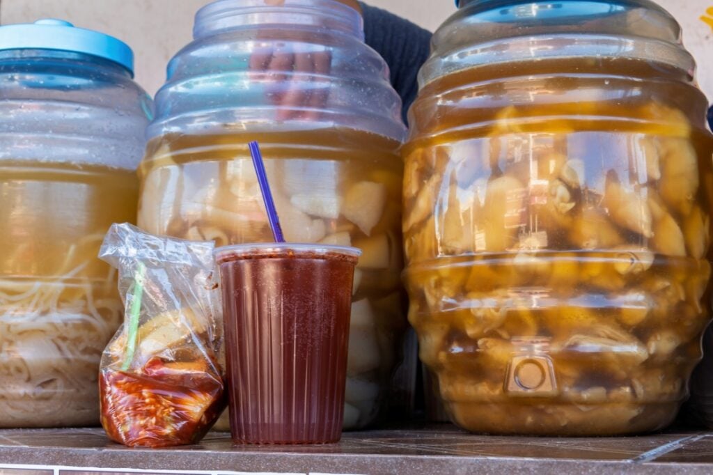 Large plastic jars filled with pickled fruits are displayed behind a plastic cup with a blue straw containing a brown liquid and a small plastic bag with red pickled vegetables and chili.