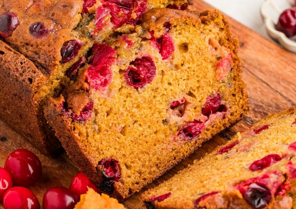 A close-up of sliced cranberry bread on a wooden board, showing moist texture with whole cranberries throughout. Some fresh cranberries and a bit of orange zest are nearby.