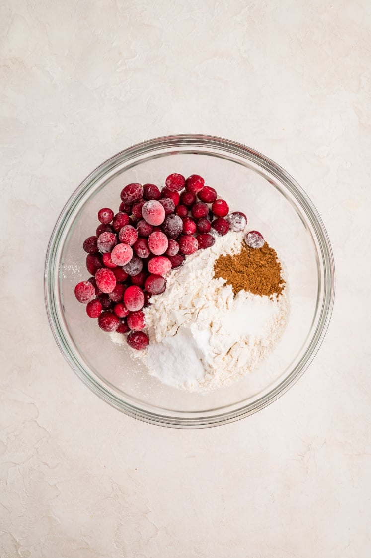 A glass bowl containing frozen cranberries, flour, baking powder, and ground cinnamon sits on a light-colored textured surface.