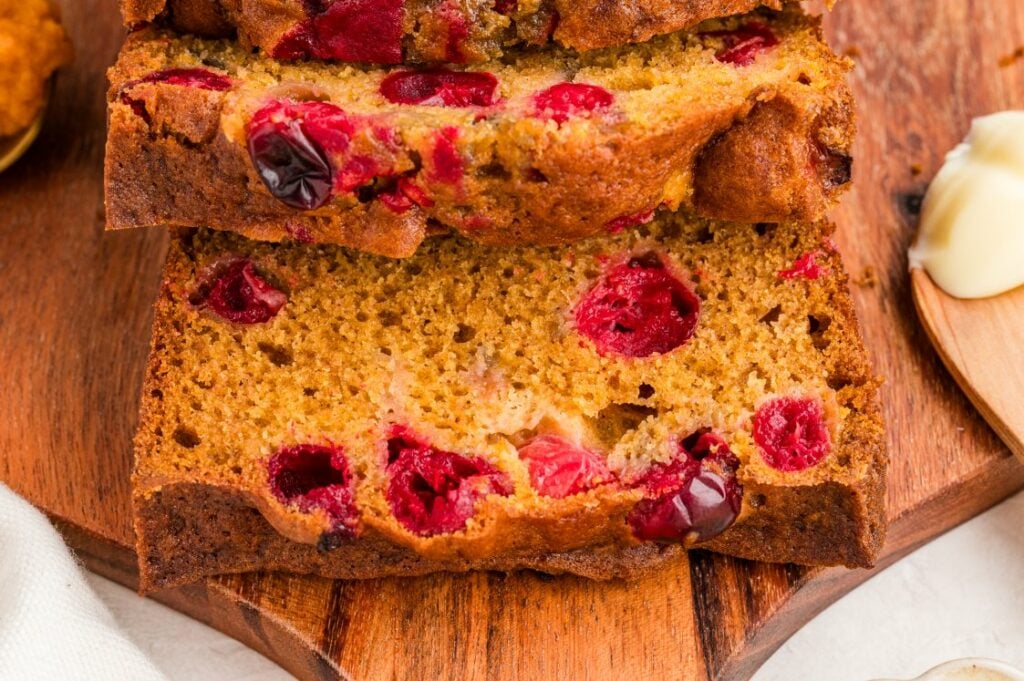 Close-up of two slices of golden-brown cranberry bread on a wooden board, showing bright red cranberries inside. A small wooden spoon with butter is visible on the right side.