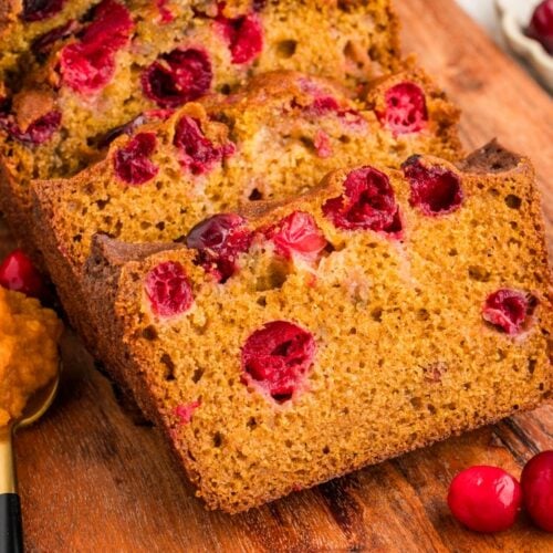 Sliced cranberry pumpkin bread on a wooden board, showing golden-brown, moist texture with visible red cranberries. A spoon with pumpkin puree and cinnamon sticks are nearby.