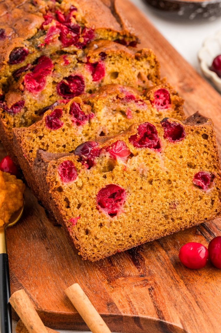 Sliced cranberry pumpkin bread on a wooden board, showing golden-brown, moist texture with visible red cranberries. A spoon with pumpkin puree and cinnamon sticks are nearby.