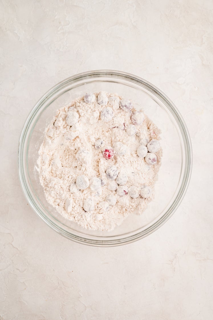 A glass bowl filled with flour and several cranberries, some of which are coated in flour, sits on a light-colored surface.