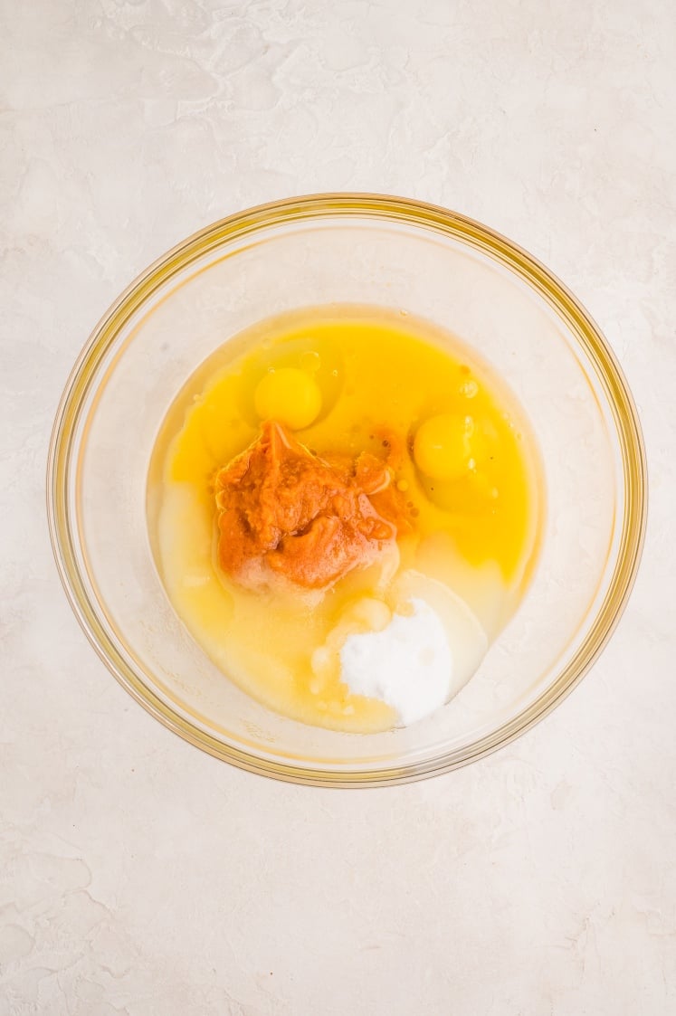 A glass mixing bowl containing two eggs, pumpkin puree, sugar, and baking powder on a light-colored countertop.