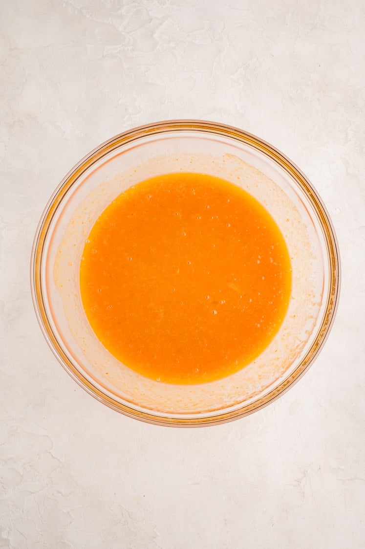 A glass bowl filled with an orange liquid mixture sits on a light-colored, textured surface.