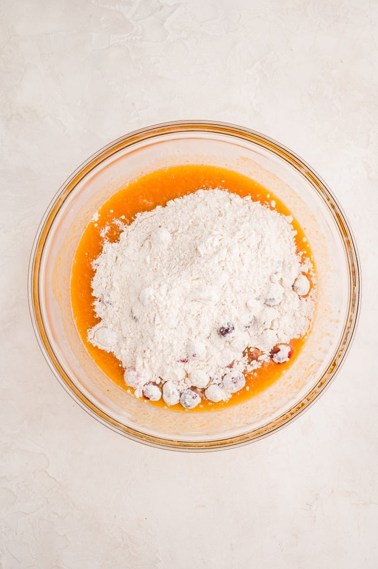 A glass mixing bowl filled with orange liquid ingredients, white flour, and blueberries sits on a light-colored countertop, ready to be mixed for baking.