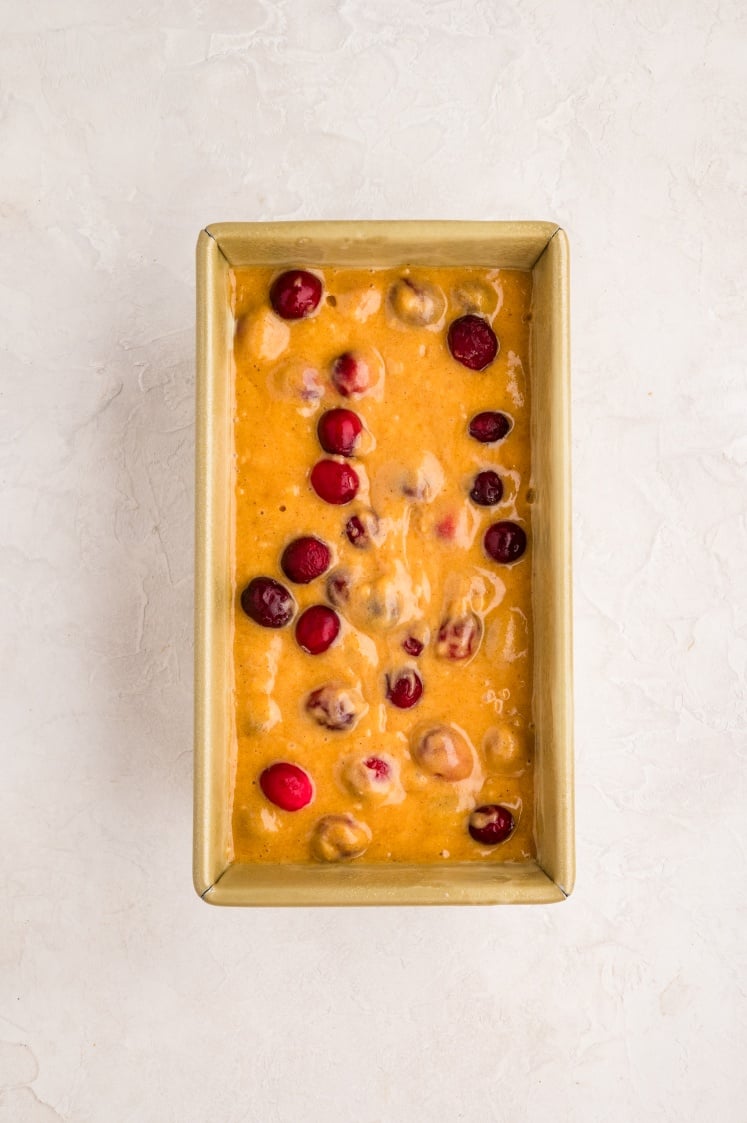 A rectangular loaf pan filled with yellow-orange batter and dotted with whole cranberries, placed on a light, textured surface.