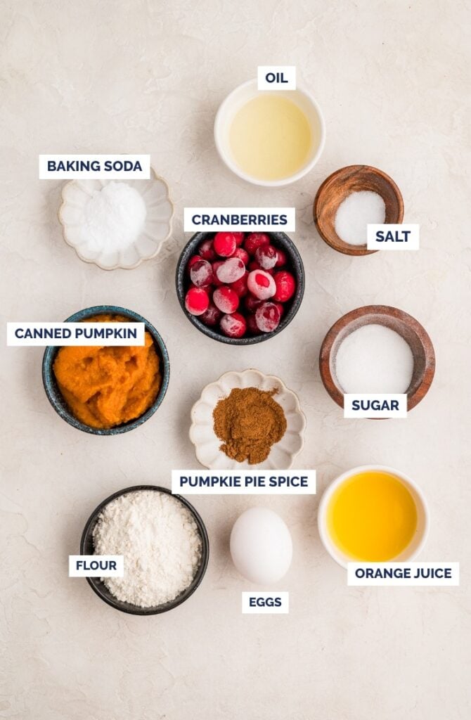 Flat lay of labeled ingredients in bowls for baking: oil, salt, sugar, orange juice, eggs, flour, pumpkin pie spice, canned pumpkin, baking soda, and cranberries on a light surface.