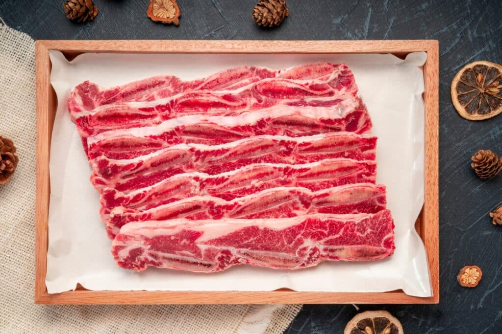 Sliced raw beef short ribs arranged neatly in a wooden tray lined with parchment paper, set on a dark surface with pinecones and dried citrus slices as decoration.