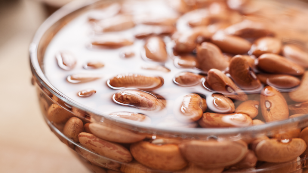 Bowl of dried pinto beans soaking in water, shown close-up with reflections on the surface.