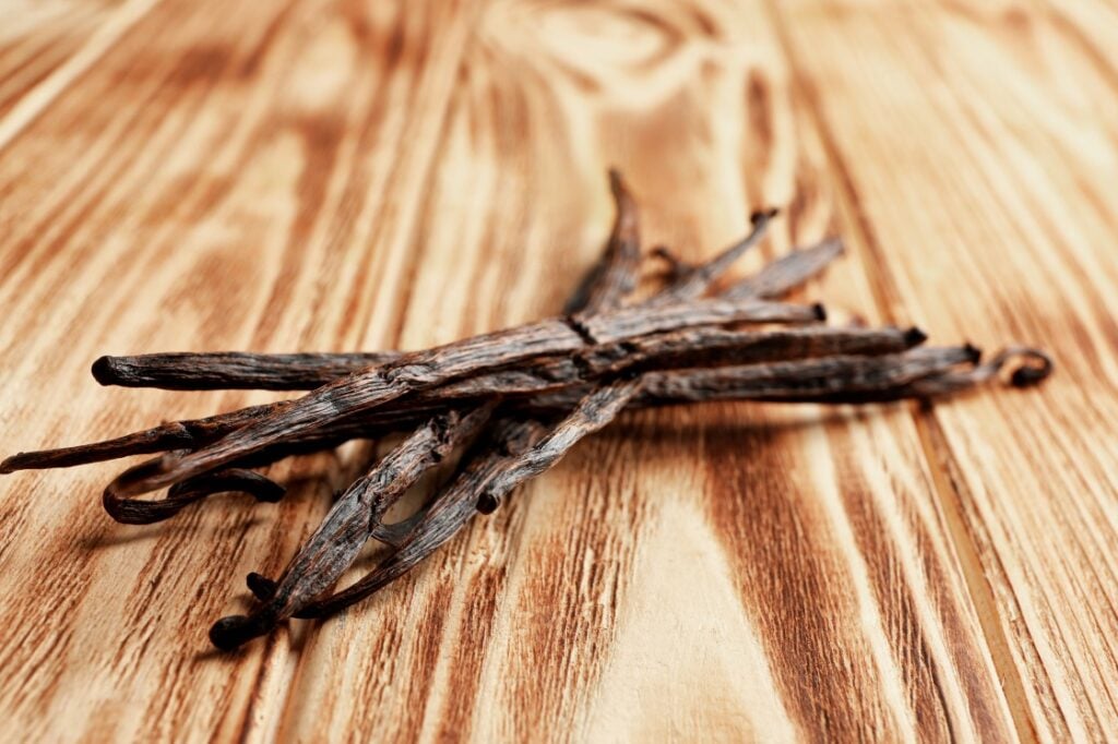 Several dried vanilla beans are arranged in a crisscross pattern on a light brown wooden surface with a natural grain pattern.