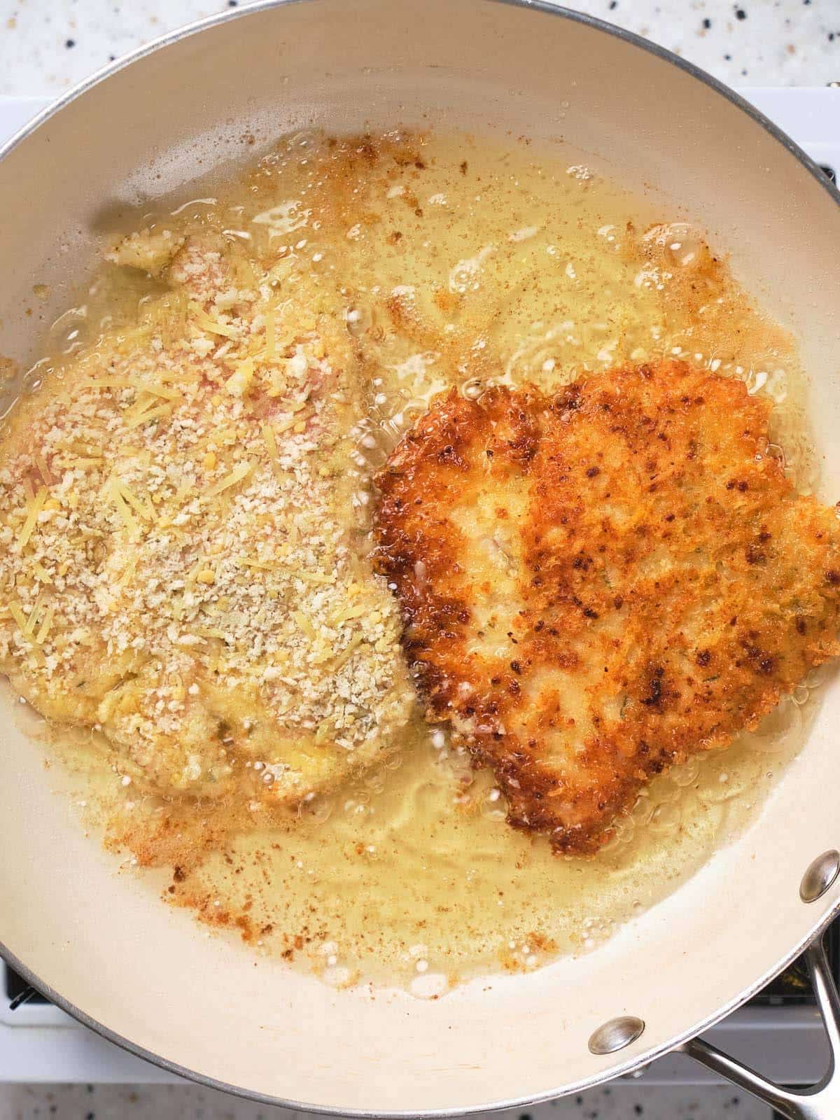Two breaded chicken cutlets frying in hot oil in a light-colored skillet; one cutlet is golden brown and crispy, while the other is still pale and partially cooked.