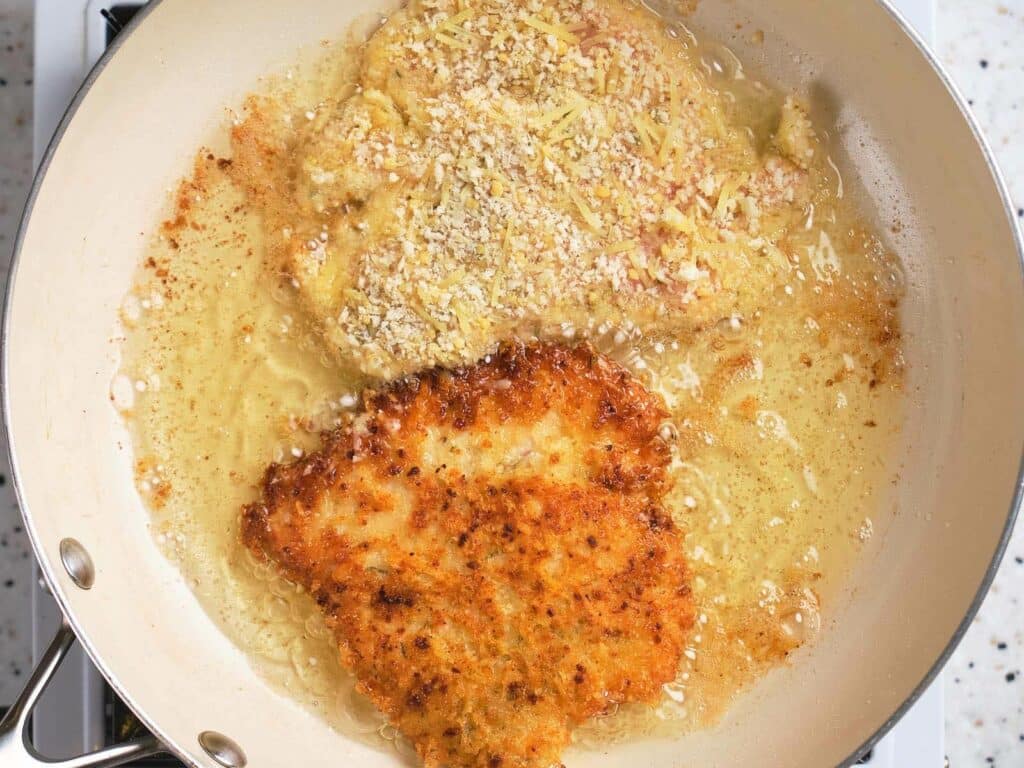 A frying pan with two breaded chicken cutlets; one is golden brown and crispy, while the other is still pale and cooking in hot oil.