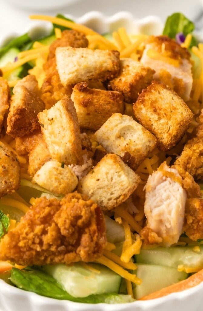 A close-up of a salad topped with crispy breaded chicken pieces, golden Air Fryer Croutons, shredded cheddar cheese, and sliced cucumbers on a bed of fresh greens.