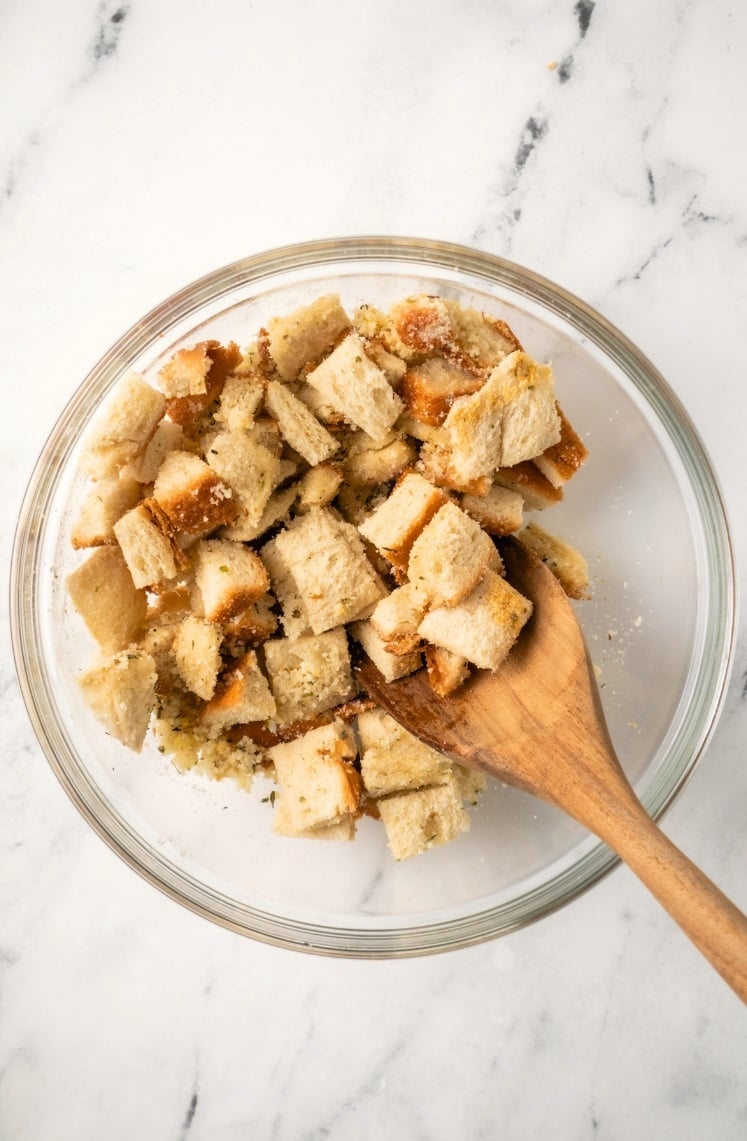 A glass bowl filled with cubed bread pieces, being mixed with a wooden spoon on a white marble surface, perfect for making homemade Air Fryer Croutons.