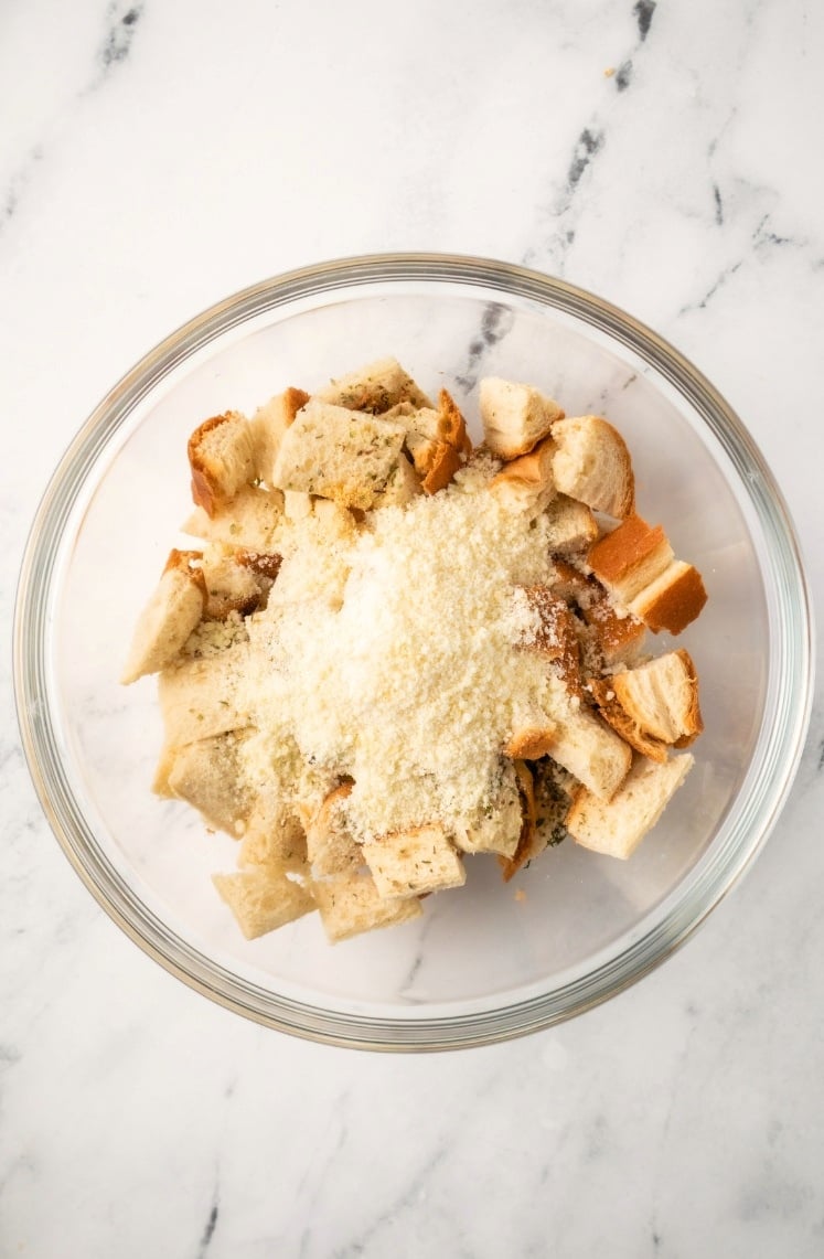 A glass bowl on a marble surface filled with cubed bread pieces, grated cheese, and seasoning, ready to be mixed for delicious homemade Air Fryer Croutons.