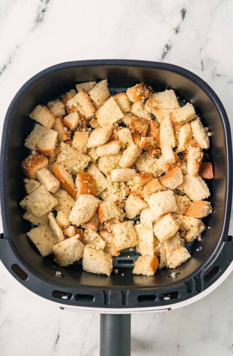 Air Fryer Croutons: Cubed pieces of bread seasoned with herbs and spices sit in an air fryer basket, ready to be toasted. The background features a clean white marble surface.