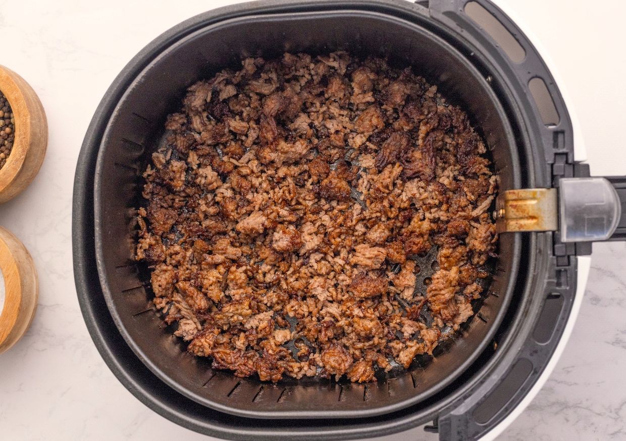 Cooked, crumbled ground meat sits inside the basket of an air fryer on a white countertop. Two small wooden bowls with spices are partially visible to the side.