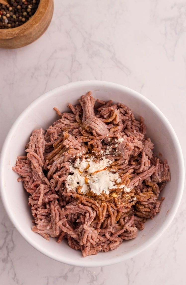 A white bowl filled with raw ground meat, topped with white flour and a brown sauce, sits on a light marble surface. A wooden bowl containing peppercorns is partially visible in the top left corner.