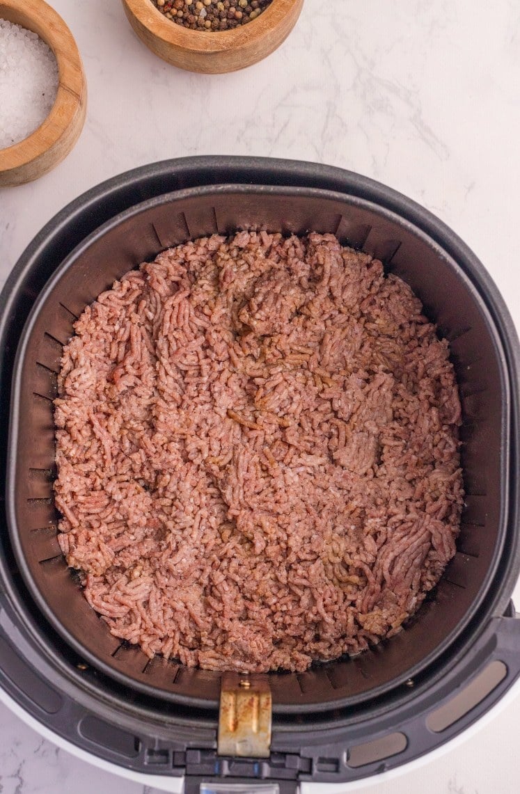 Raw ground beef in an air fryer basket, ready to be cooked. Small wooden bowls of salt and peppercorns are visible on a white marble countertop in the background.